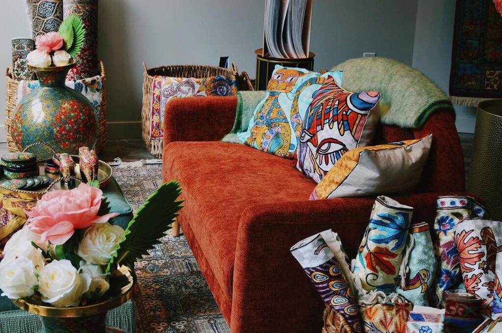 A vibrant room with a red armchair, embroidered Kashmiri cushions, handwoven rugs, floral decor, and artisanal crafts on display.